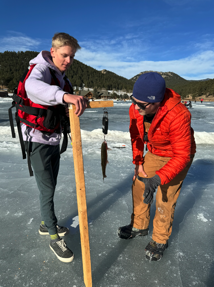 Two people on ice with a freshly caught fish hanging from a wooden stand.