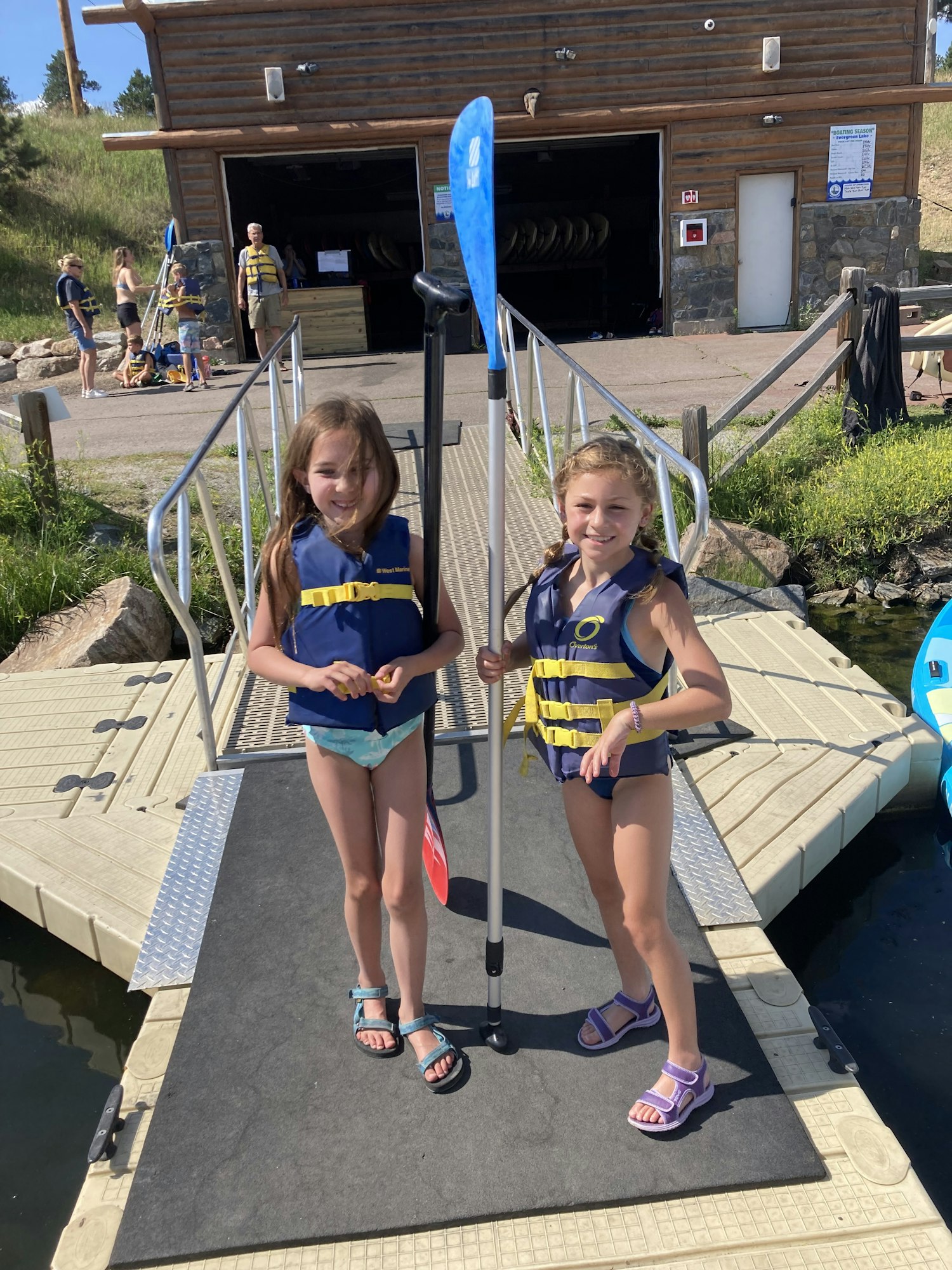 Two kids in life jackets holding paddles, standing on a dock near the water, ready for a water activity.