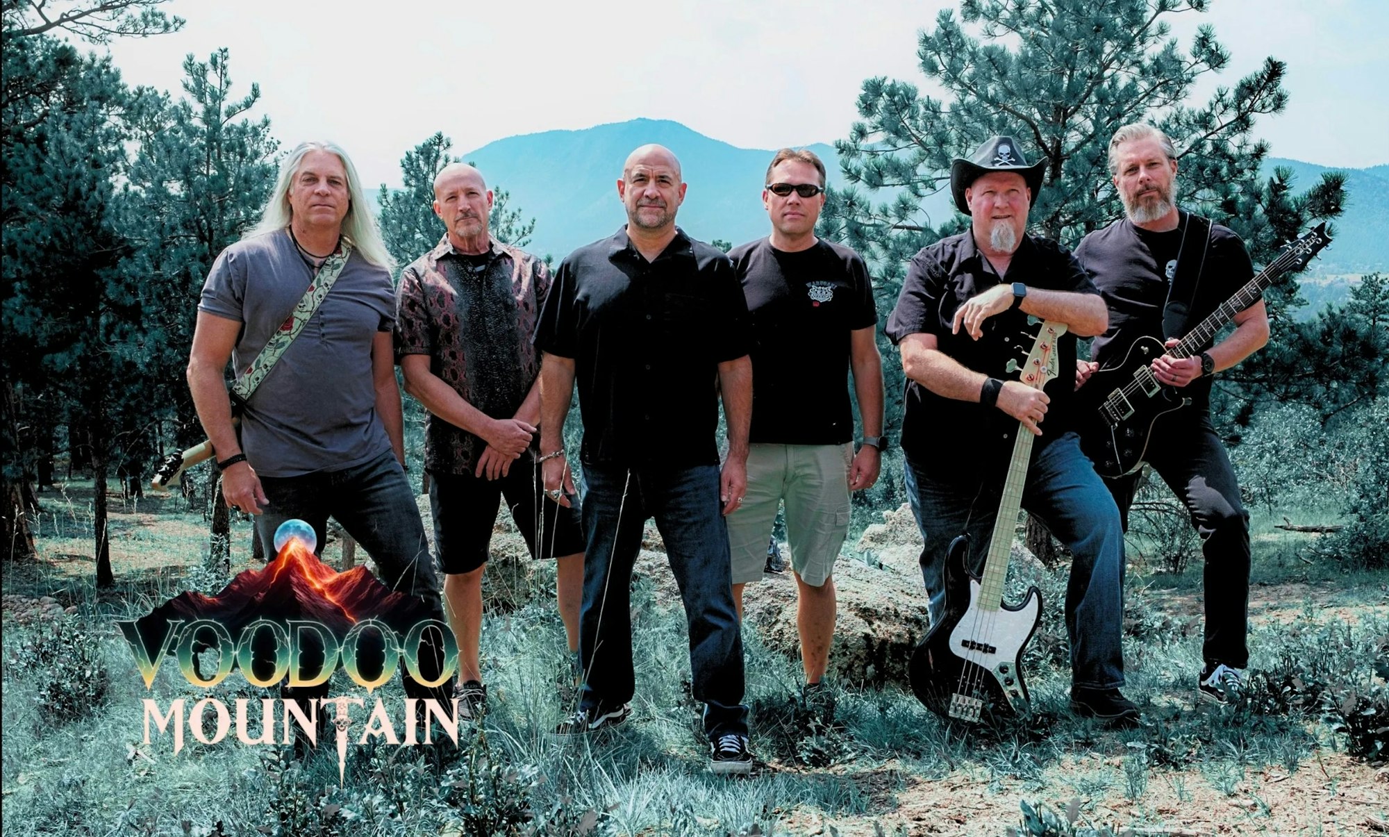 A group of six musicians stands together outdoors, surrounded by trees and mountains, promoting "Voodoo Mountain."