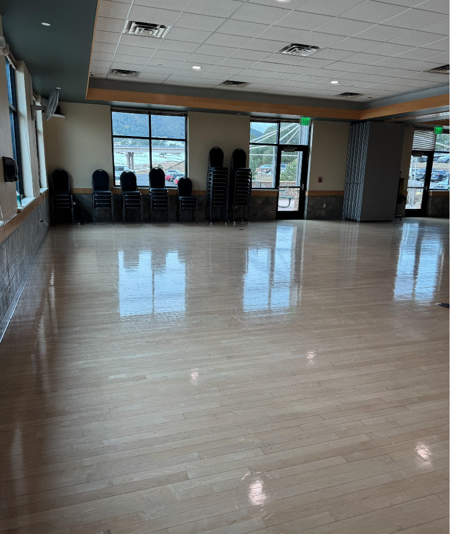 An empty room with a wooden floor, stacked chairs, and large windows.