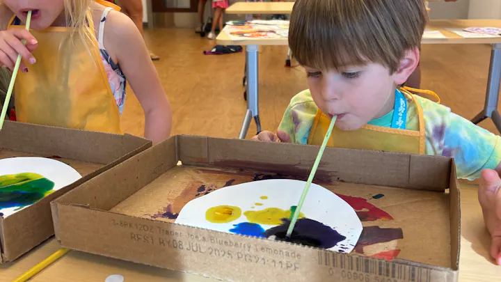 Children creating art by blowing paint with straws on paper inside cardboard trays.