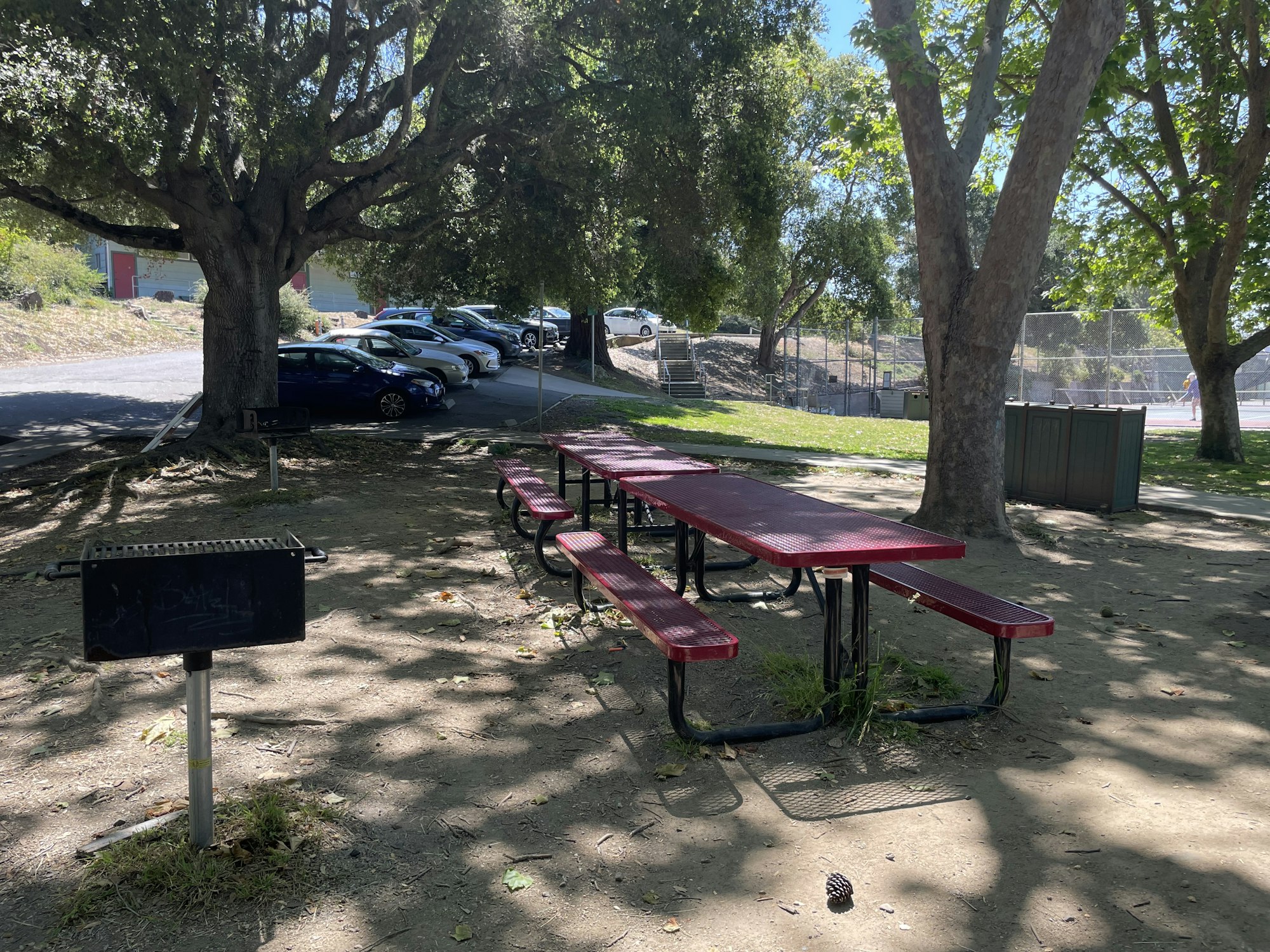 A picnic area with red tables, a grill, and trees, with parked cars and a tennis court in the background.