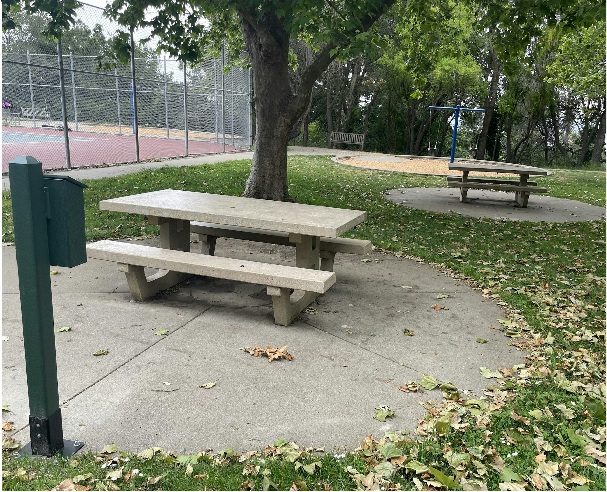 A park with picnic tables, a swing set, a tennis court, and scattered autumn leaves.