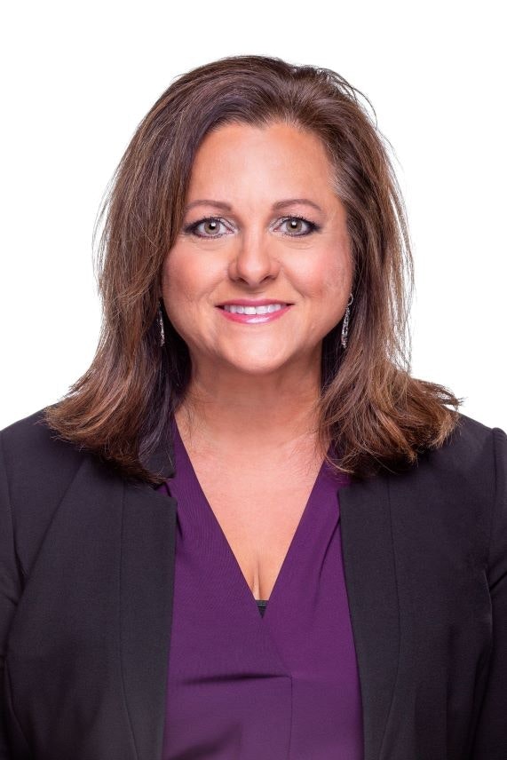 A professional portrait of a woman with medium brown hair, wearing a black blazer and a purple top, smiling against a white background.