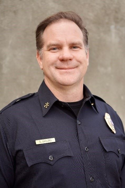 A smiling man in a police uniform with a badge stands against a neutral background.