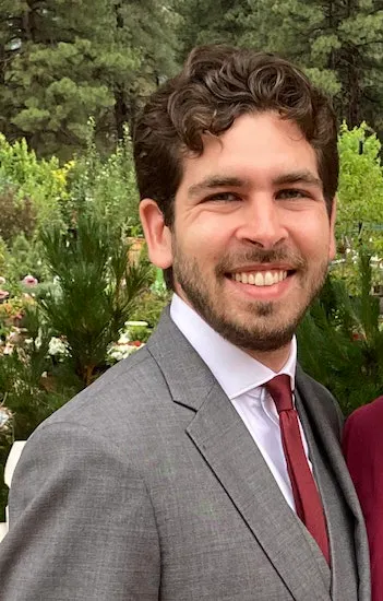 A man in a suit smiles warmly against a backdrop of greenery, conveying a pleasant and cheerful atmosphere.