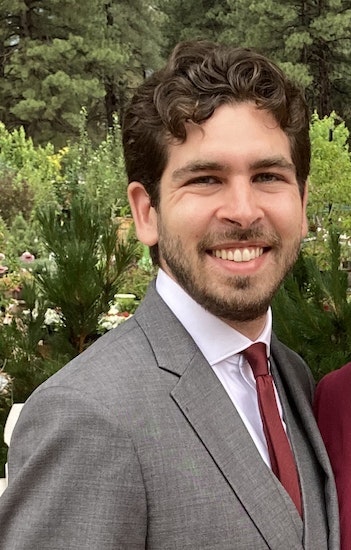 A man in a suit smiles warmly against a backdrop of greenery, conveying a pleasant and cheerful atmosphere.