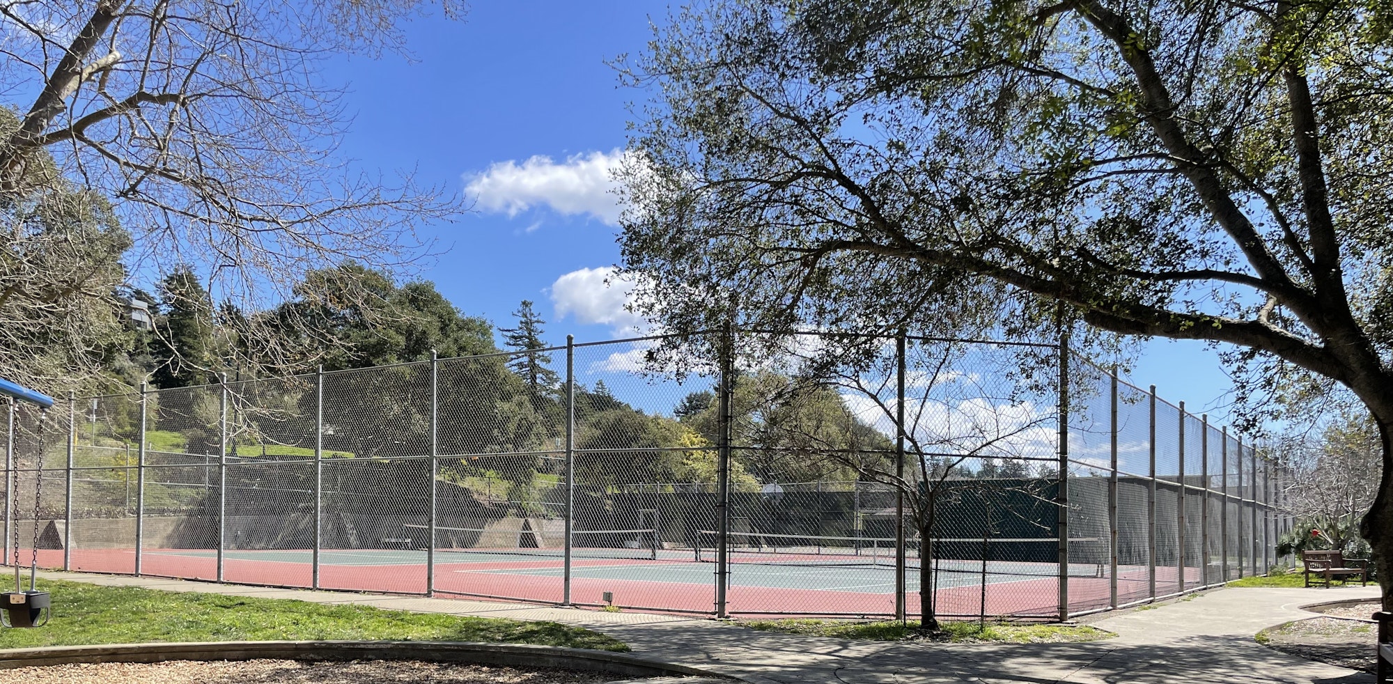 A fenced tennis court in a park with trees, swings, and a clear blue sky.