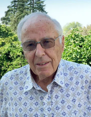 An elderly man wearing glasses and a patterned shirt stands in front of greenery on a sunny day.