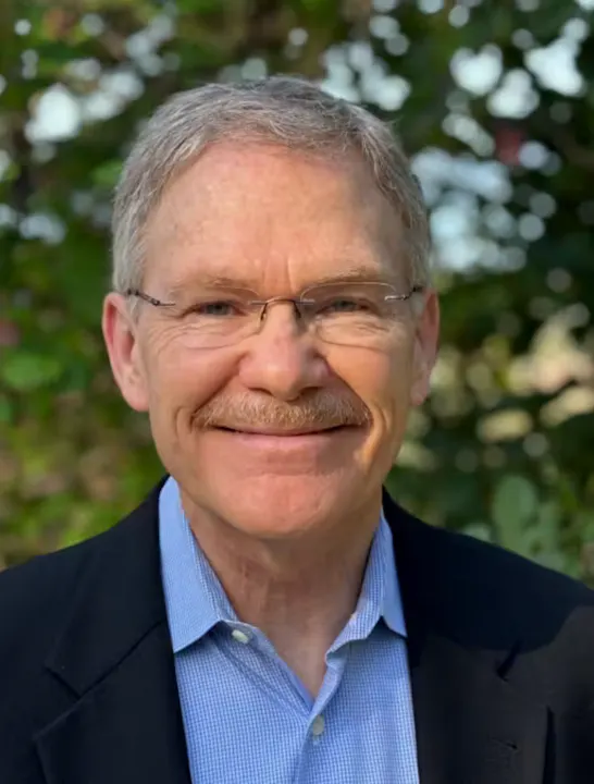The image shows a smiling man with glasses and a mustache, wearing a blue shirt and a black jacket against a green background.