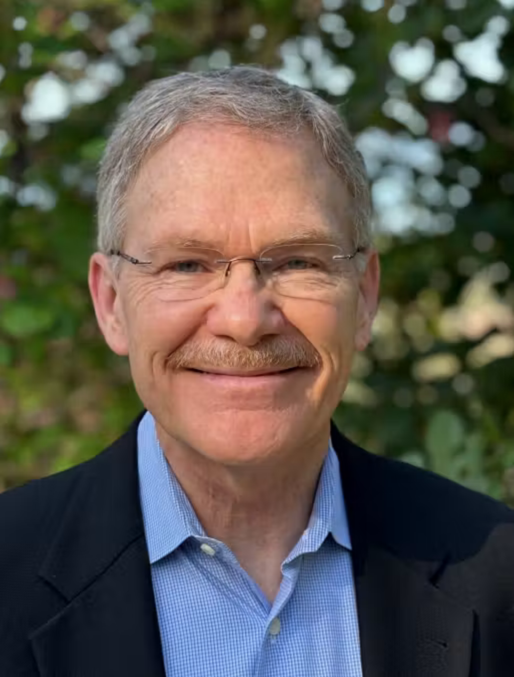 The image shows a smiling man with glasses and a mustache, wearing a blue shirt and a black jacket against a green background.