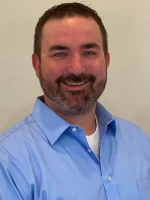 The image shows a smiling man with a beard, wearing a blue dress shirt, set against a light background.