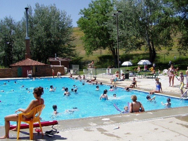 People swimming and relaxing around an outdoor pool on a sunny day, with trees and grass in the background.