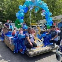 A parade float decorated with blue balloons, individuals seated, and two people sitting upfront with a balloon arch overhead.