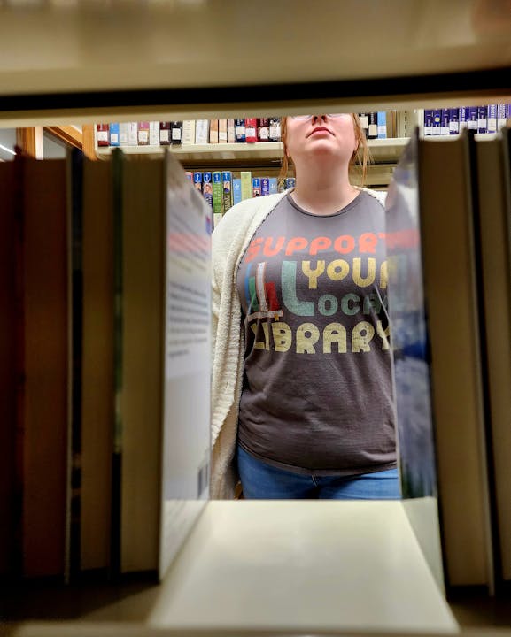 Person in a library seen through a book gap, wearing a shirt that says "Support Your Local Library."