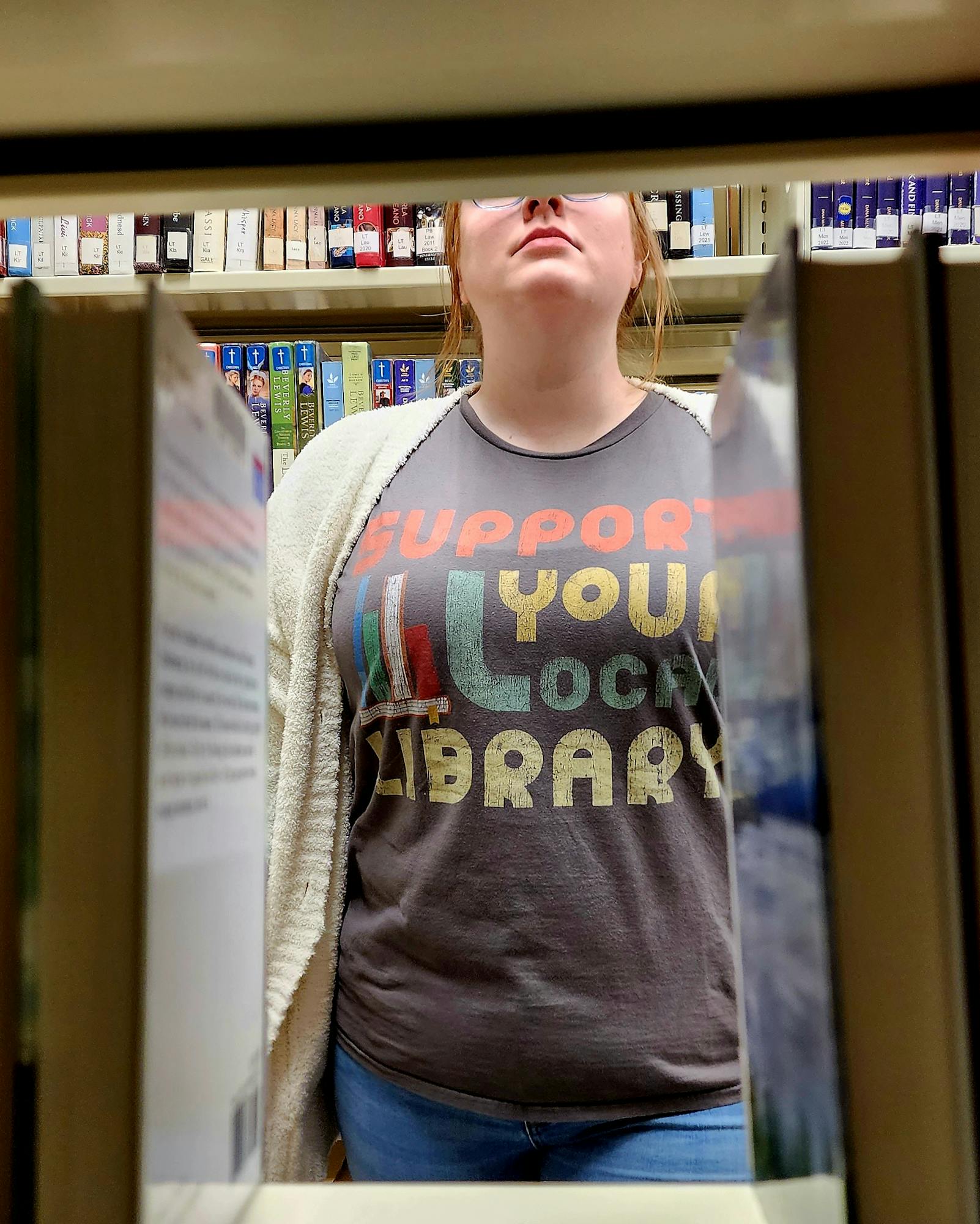 Person in a library seen through a book gap, wearing a shirt that says "Support Your Local Library."