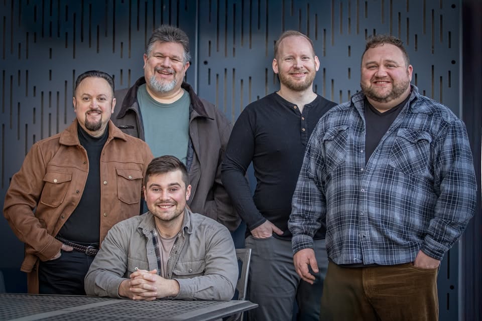 Five men posing together, with one seated and four standing behind, against a patterned backdrop.