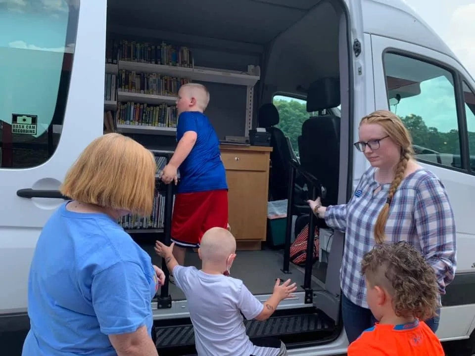 A mobile library van with books, a librarian, and children exploring the van's collection.