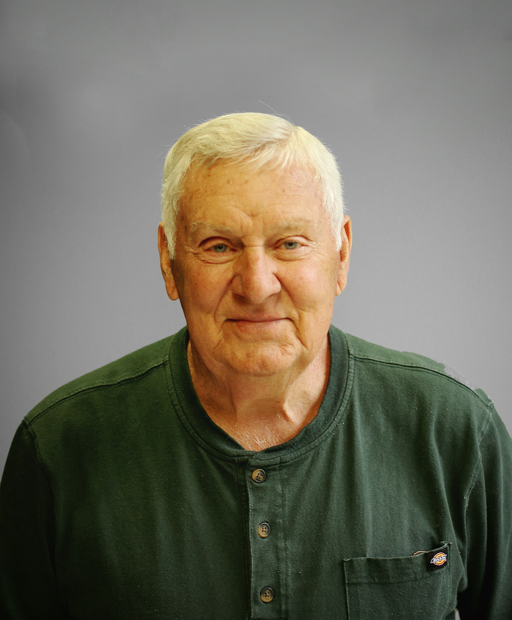 Elderly man with white hair wearing a green buttoned shirt, against a plain background.