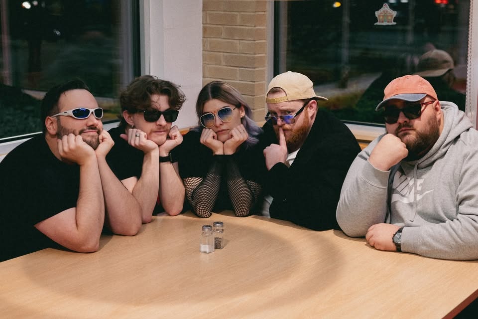 Five people wearing sunglasses pose with their hands under their chins, sitting around a table with salt and pepper shakers.