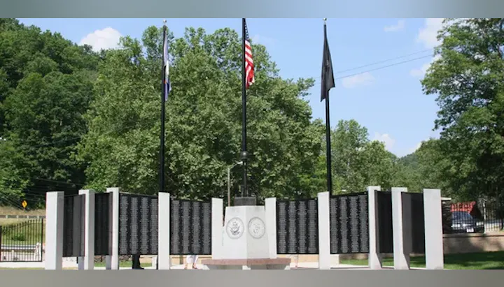 Military memorial with flags and engraved black panels, surrounded by white pillars under a clear sky.