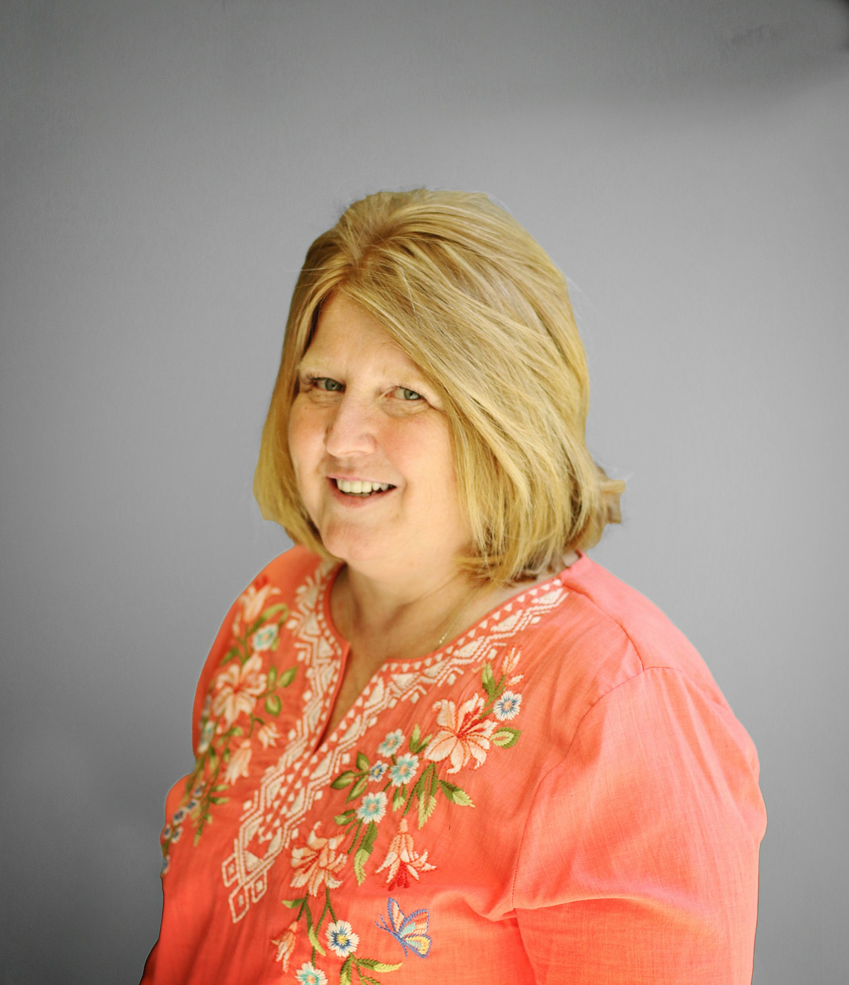 A person with shoulder-length hair wearing a coral top with floral embroidery, smiling against a gray background.