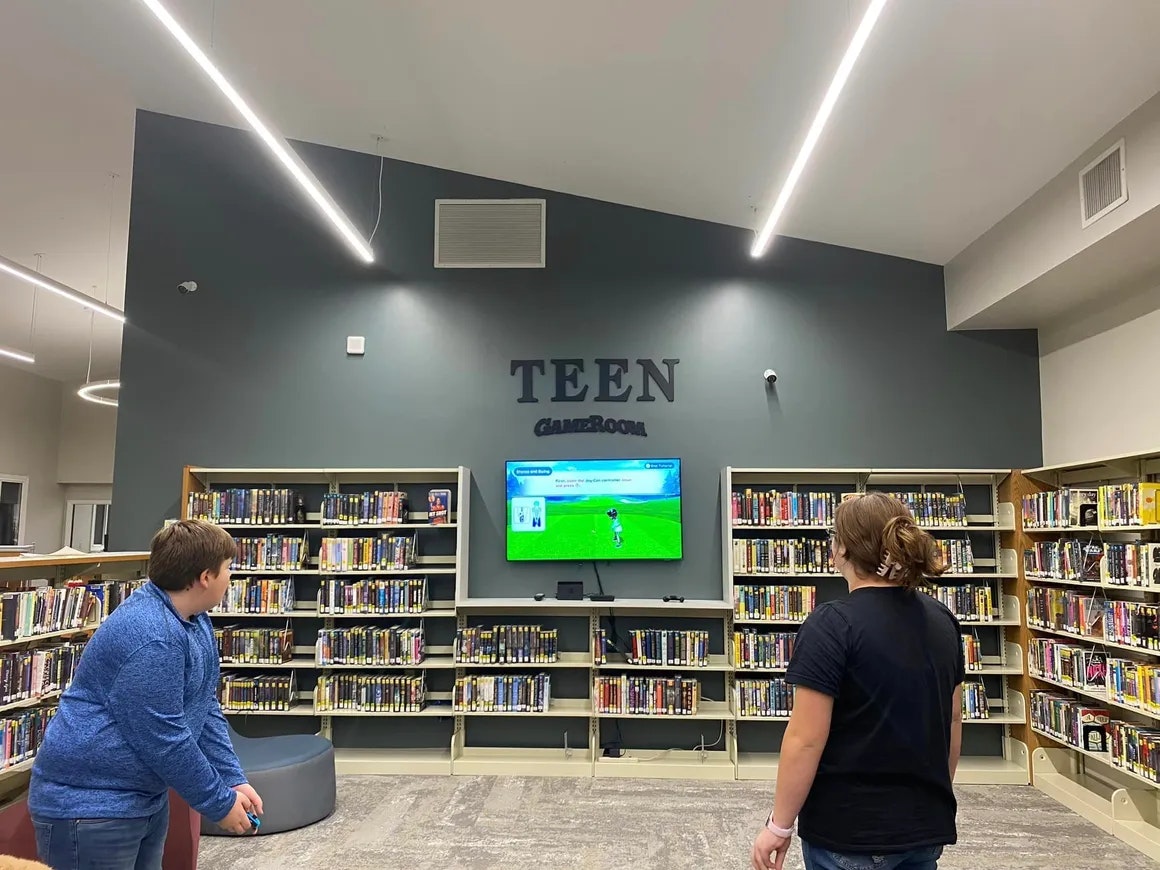 Two individuals playing a video game in a library's designated 'TEEN GameRoom' area, surrounded by bookshelves.