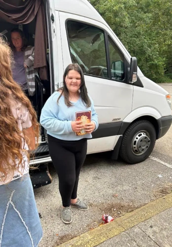 A person standing in front of a van smiling, holding a book, with another person inside the van and an individual's partial view in the foreground.