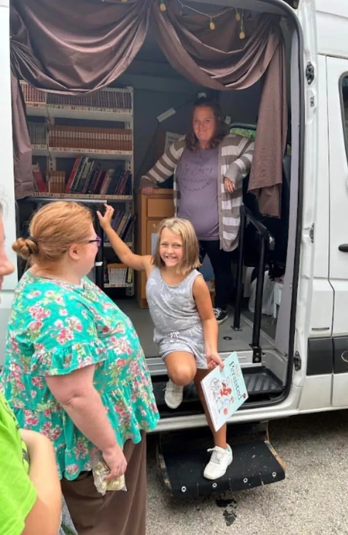 A mobile library van with shelves of books, a smiling child exiting holding a book, and an adult gesturing towards her.