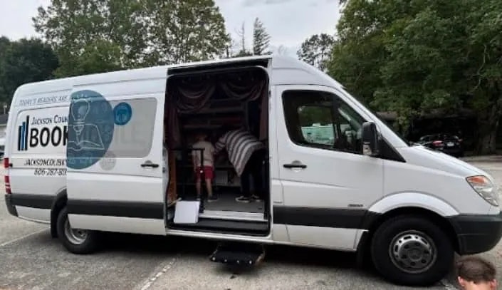 A van with an open side door revealing shelves of books inside, suggesting a mobile library or bookmobile service.