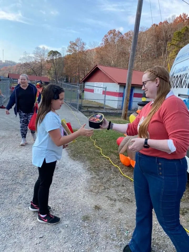 A girl is creating a giant bubble with the help of a smiling woman; they're using a bubble wand. People and autumn trees are in the background.