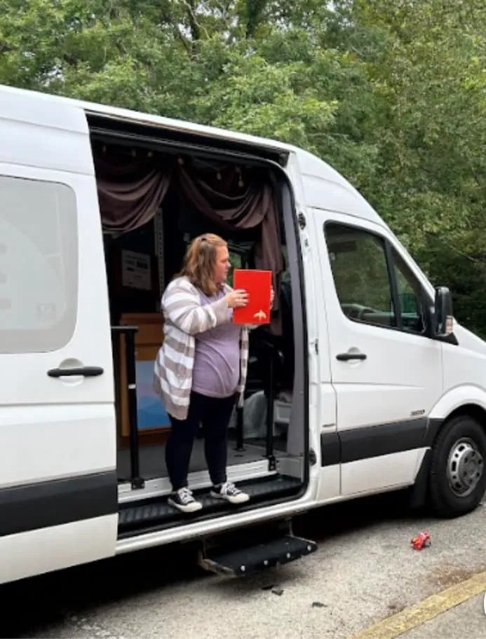 A person stands in a van doorway, holding a red book, curtains behind, surrounded by greenery.