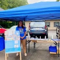 A person under a blue canopy next to a table with informational materials, outdoors.