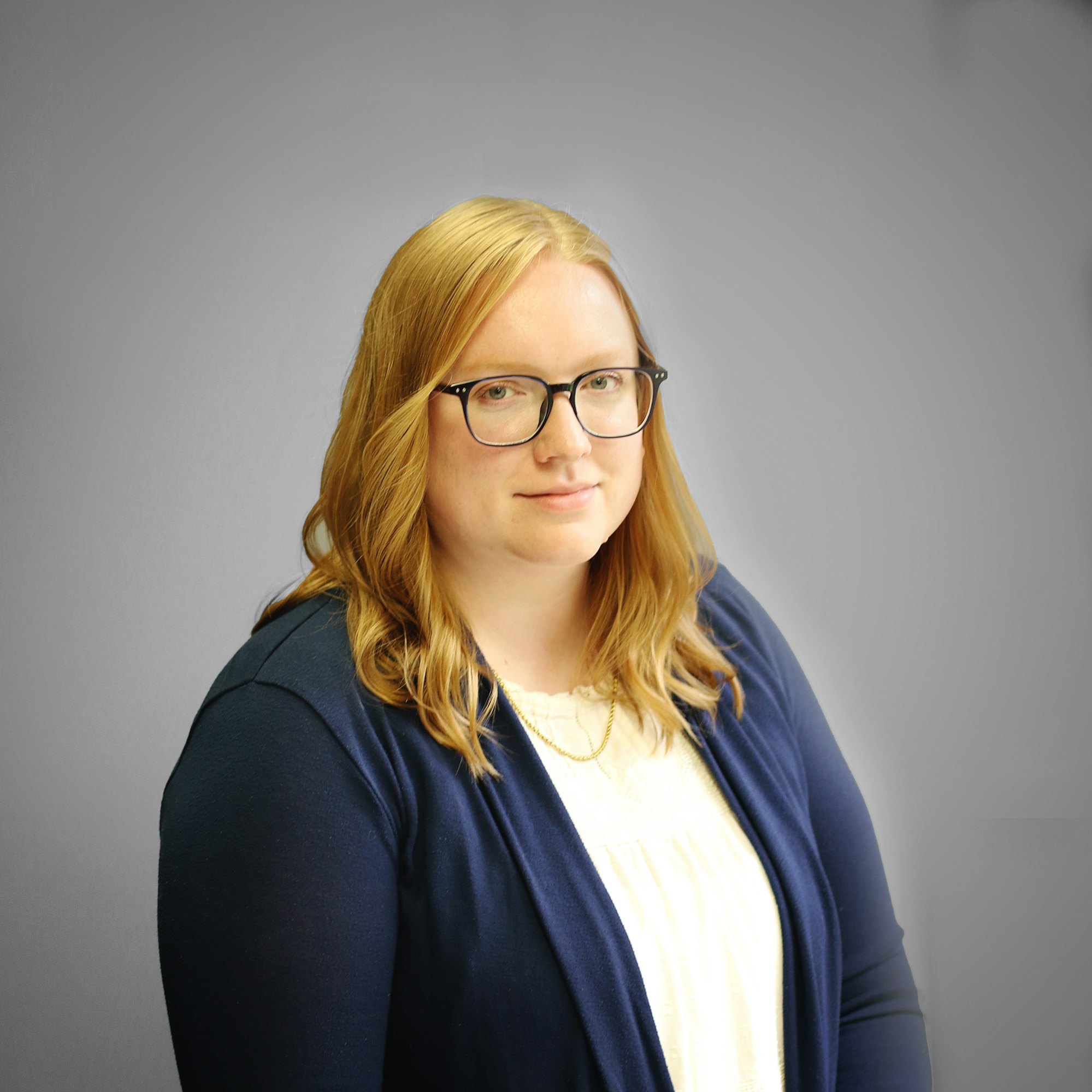 A person with glasses, wearing a white blouse and blue cardigan, against a gray background.