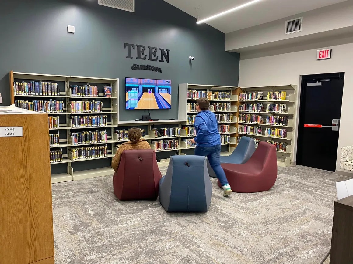 A teen game room in a library with shelves of books, modern seating, and a bowling game on TV, with two people present.