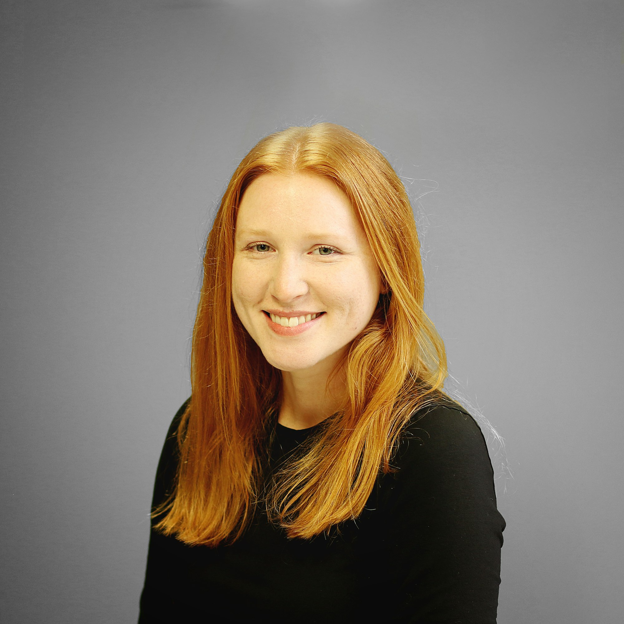 A person with long red hair smiles against a gray background, wearing a black top.