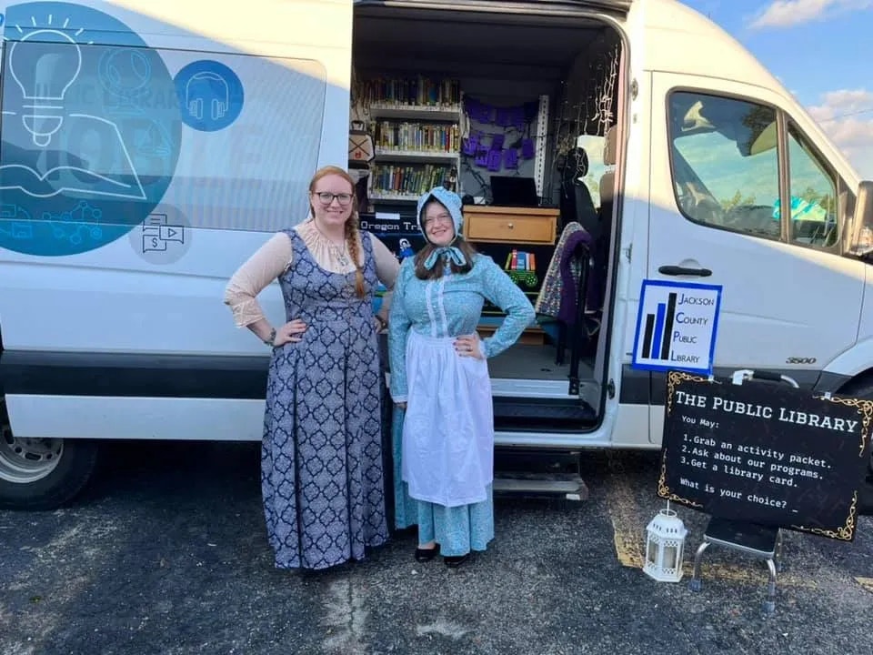 Two people standing by a van converted into a mobile library; signs promote library services.