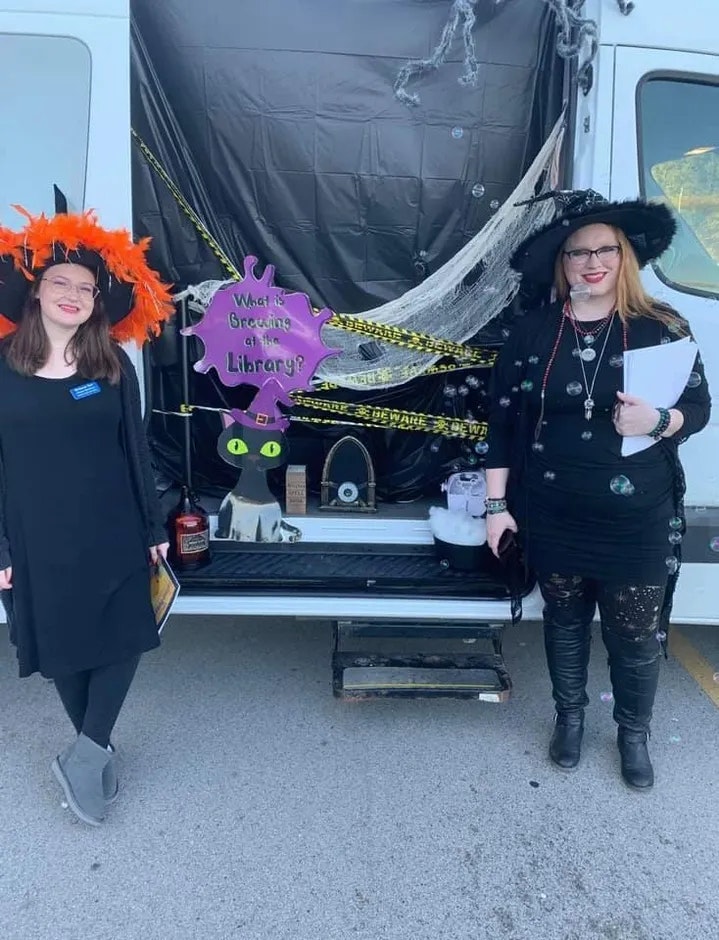 Two people in Halloween costumes standing near a decorated vehicle with a sign that says "What is Brewing at the Library?"