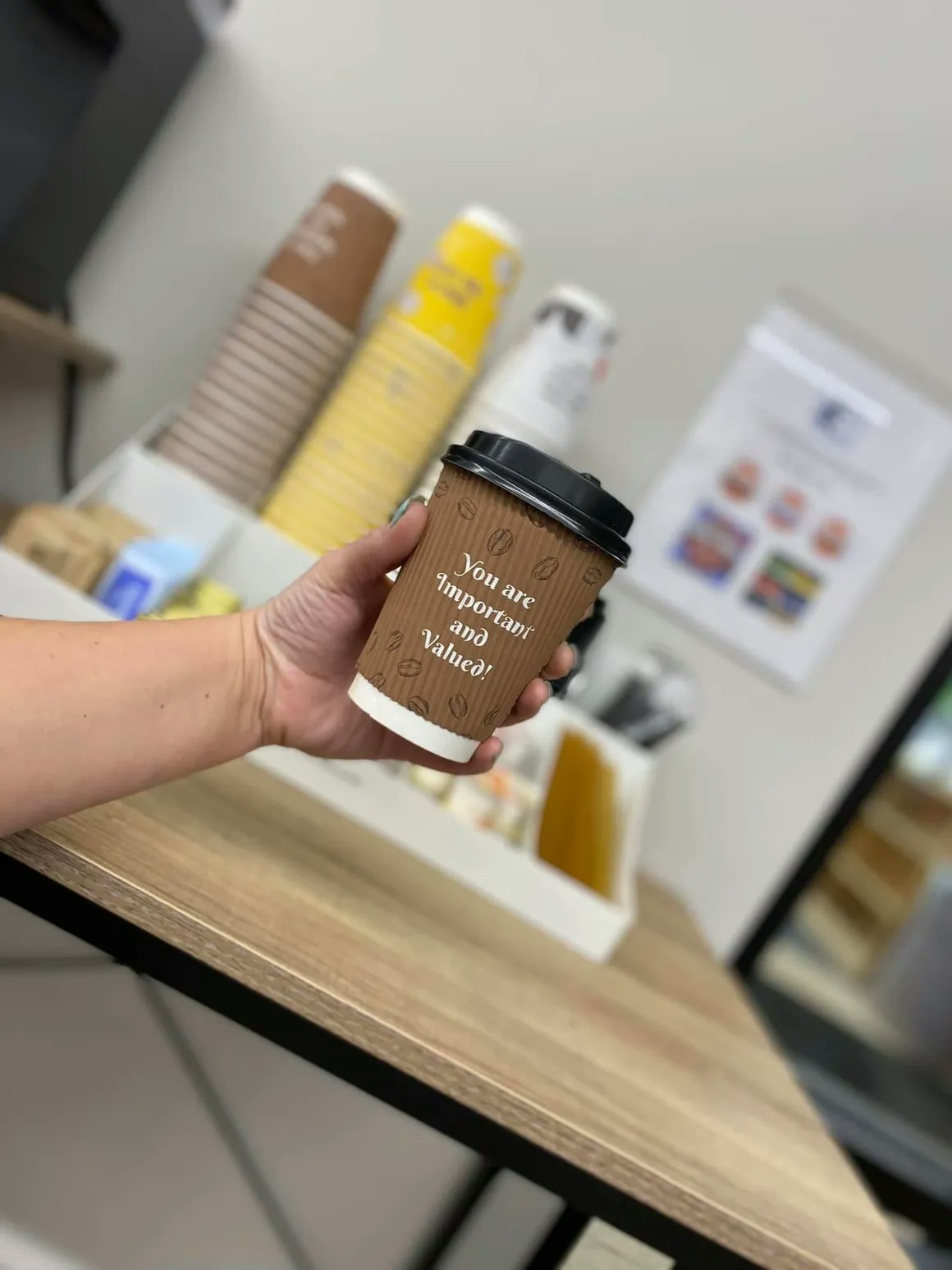 A hand holding a coffee cup with an inspirational message, blurred background of a coffee station.