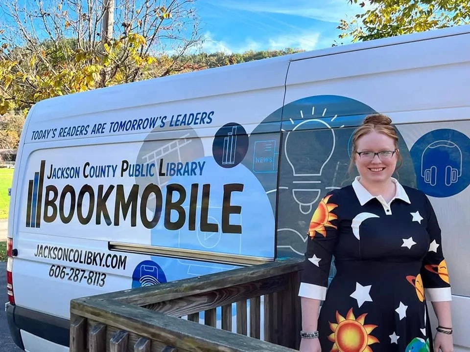 A smiling person stands beside a 'bookmobile' from the Jackson County Public Library, promoting reading and literacy.