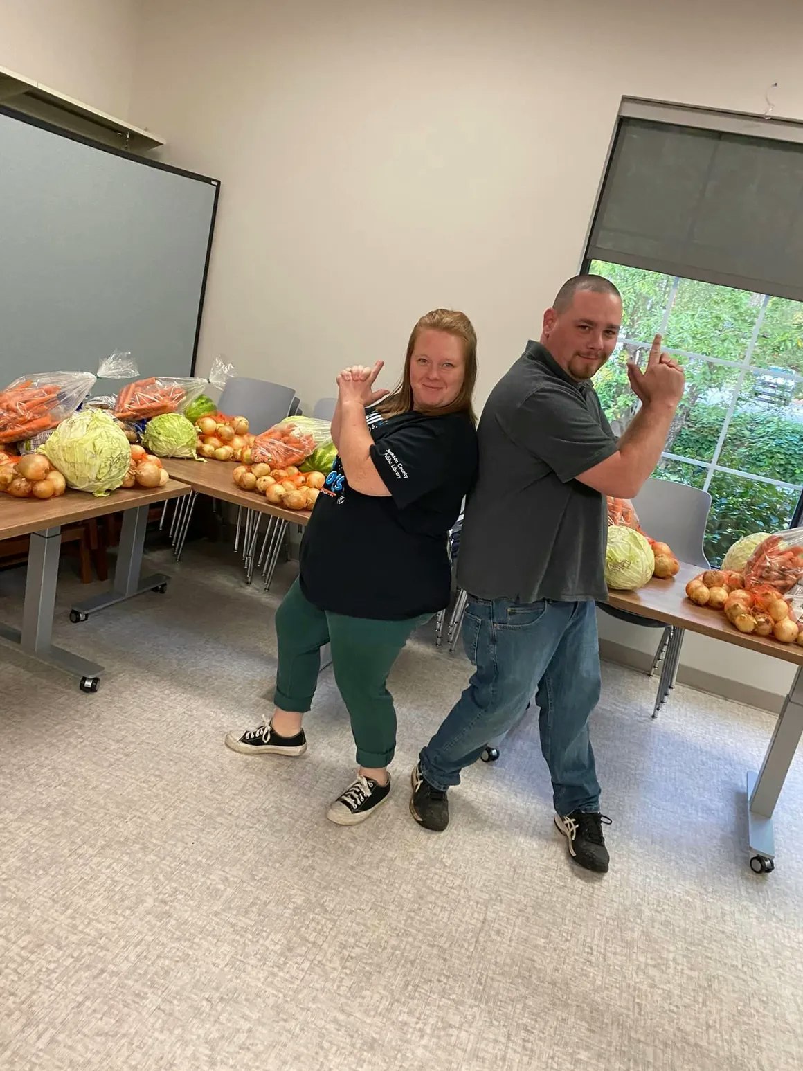Two individuals posing back-to-back with fresh produce on a table behind them.