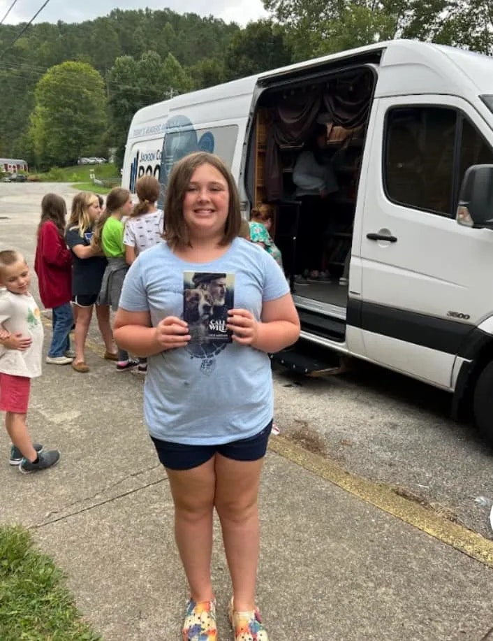 A girl stands smiling before a mobile library van, proudly holding a book. Other kids queue in the background.