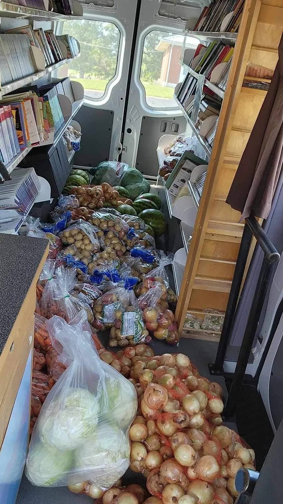 A van filled with books and fresh produce like watermelons, potatoes, and onions.