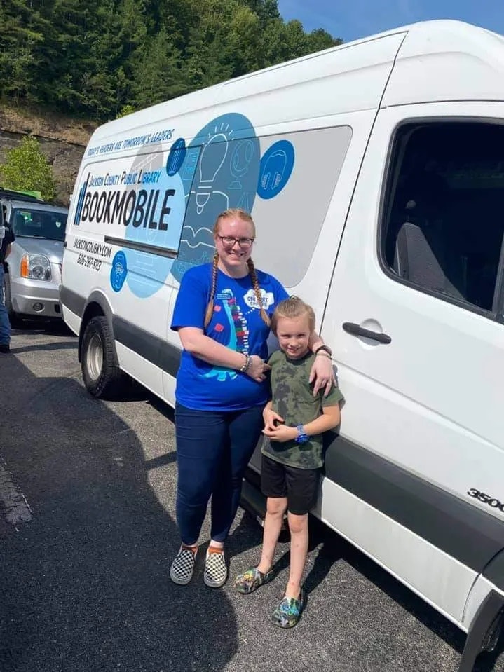 Two individuals in front of a 'Bookmobile' from a library service, standing on a sunny day.