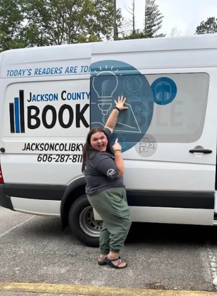 A person is playfully posing with a library bookmobile, matching their hand to a graphic hand on the vehicle.