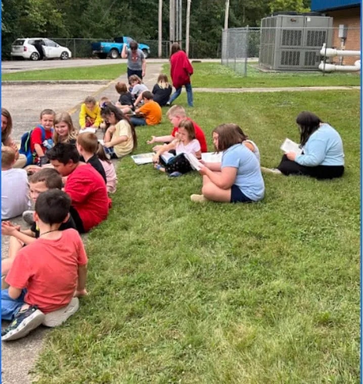 Children sitting on grass, reading and interacting, outdoors near buildings and vehicles.