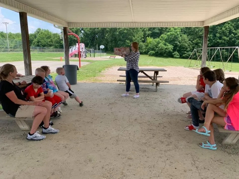 A group of children sitting, listening to a woman reading a book outdoors under a shelter. Playground in the background.
