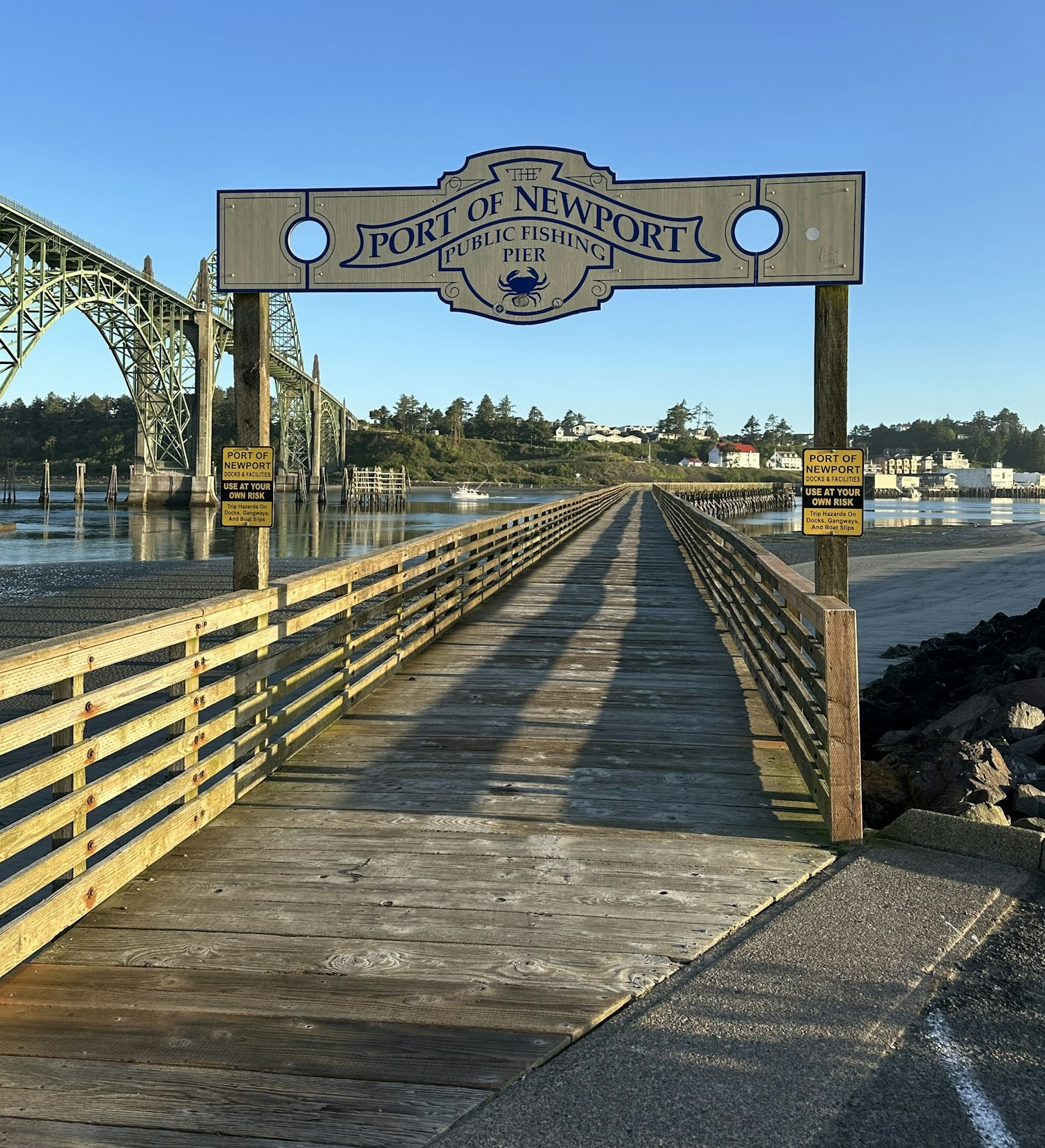 A wooden pier with "Port of Newport Public Fishing Pier" sign, bridge in the background, clear sky.