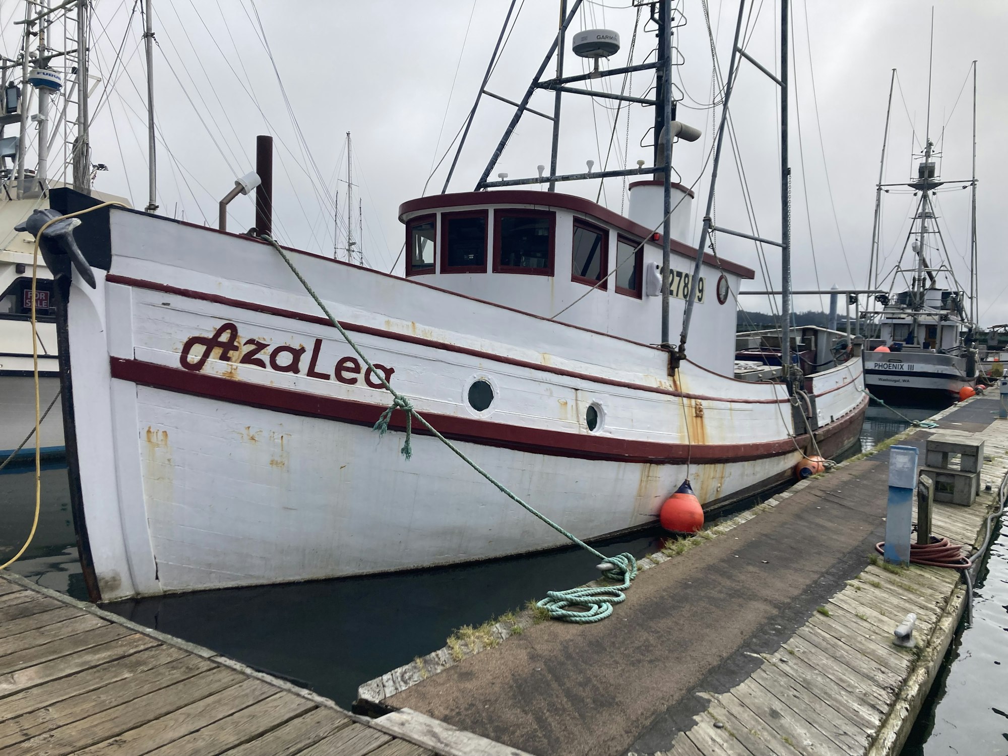 A white fishing boat named "Azalea" docked at a pier with other boats and maritime equipment in the background.