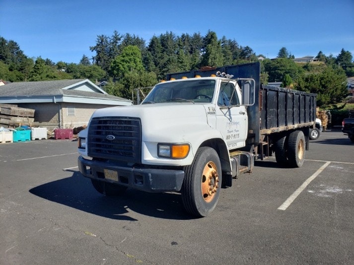 The image shows a white Ford dump truck parked in a lot with trees and buildings in the background.
